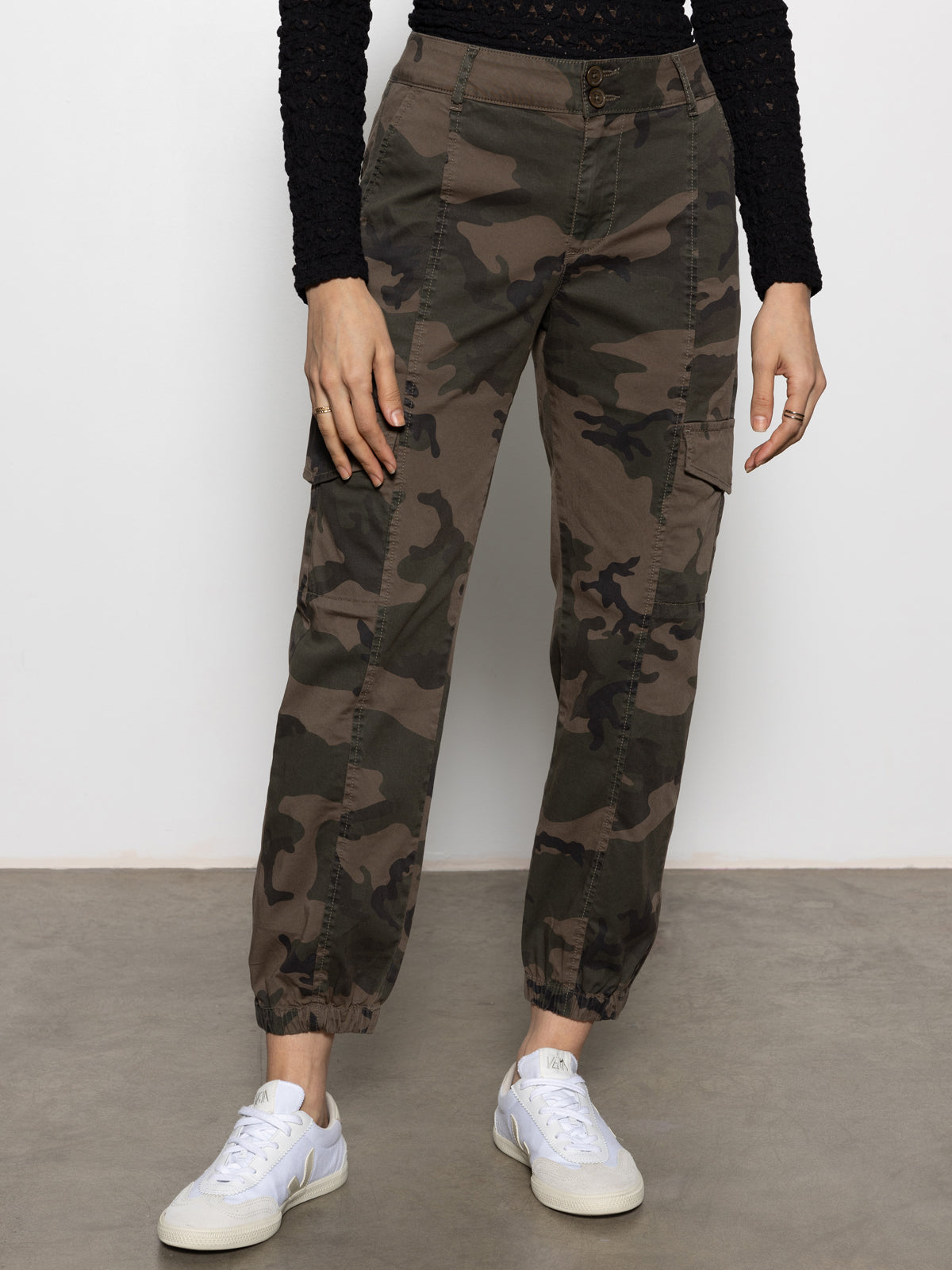 Wearing Sanctuary Clothing™s rebel pant hiker camo with a black textured long-sleeve top and white sneakers, the person stands against a plain wall, hands relaxed at their sides. Image cropped from chest down.