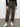 Wearing Sanctuary Clothing™s rebel pant hiker camo with a black textured long-sleeve top and white sneakers, the person stands against a plain wall, hands relaxed at their sides. Image cropped from chest down.