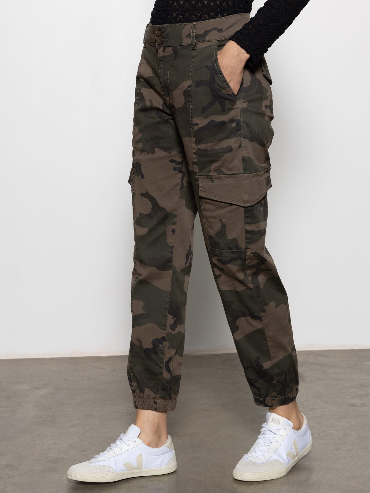 A person wears Sanctuary Clothing's rebel pant hiker camo cargo pants with elastic cuffs, a black patterned long-sleeve top, and white sneakers, standing on a gray floor against a plain white background.