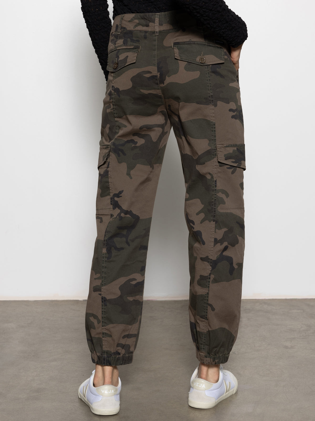 A person wearing Sanctuary Clothing rebel pant hiker camo, featuring side and back pockets with elastic ankle cuffs, and white sneakers stands facing a white wall, shown from the back.