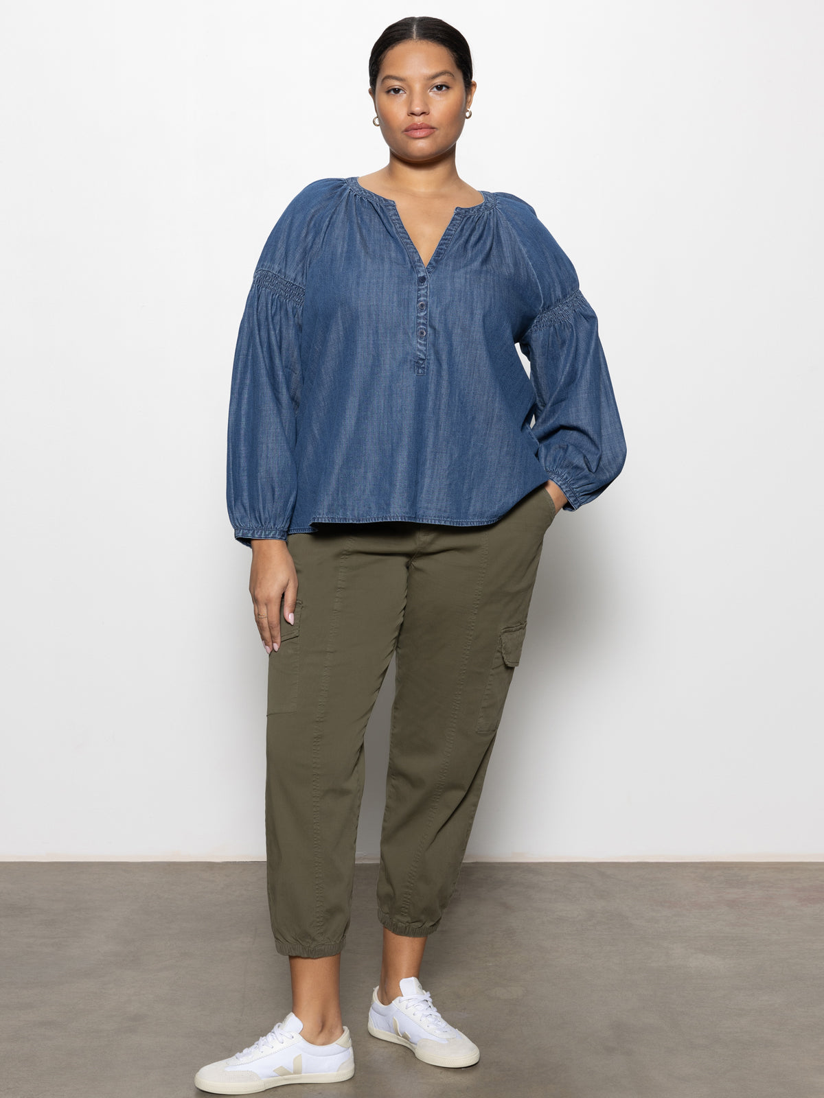 A woman stands against a white wall wearing Sanctuary Clothing™s rebel pant hiker green from the inclusive collection, paired with a loose denim blouse and white sneakers. One hand in her pocket, she maintains a neutral expression.