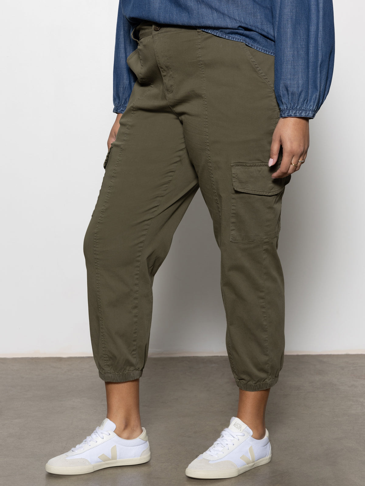 Wearing Sanctuary Clothing's rebel pant hiker green from the inclusive collection, a person stands on a gray floor against a plain white wall, showing only the lower half of their body.