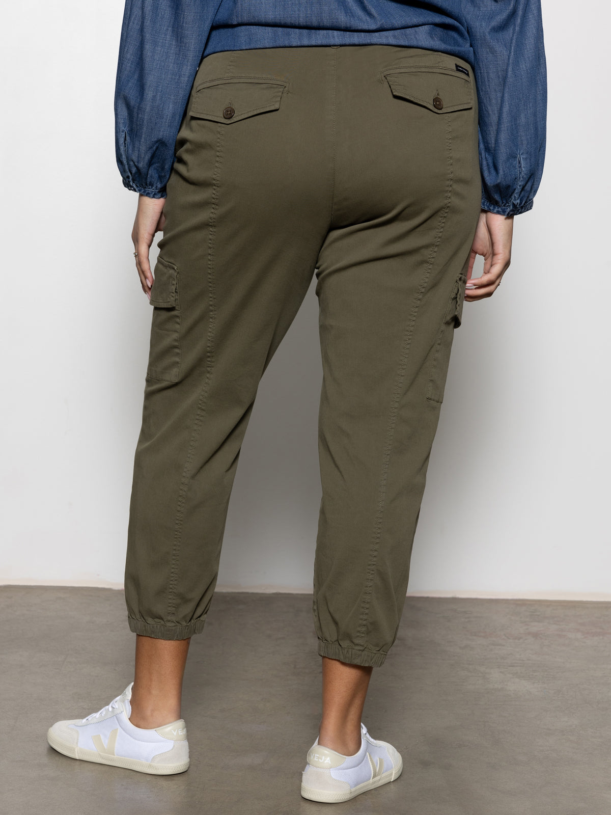 A person wearing Sanctuary Clothing's Rebel Pant Hiker Green from the Inclusive Collection, paired with white sneakers and a blue long-sleeve shirt, is shown from the back standing on a concrete floor against a light wall.