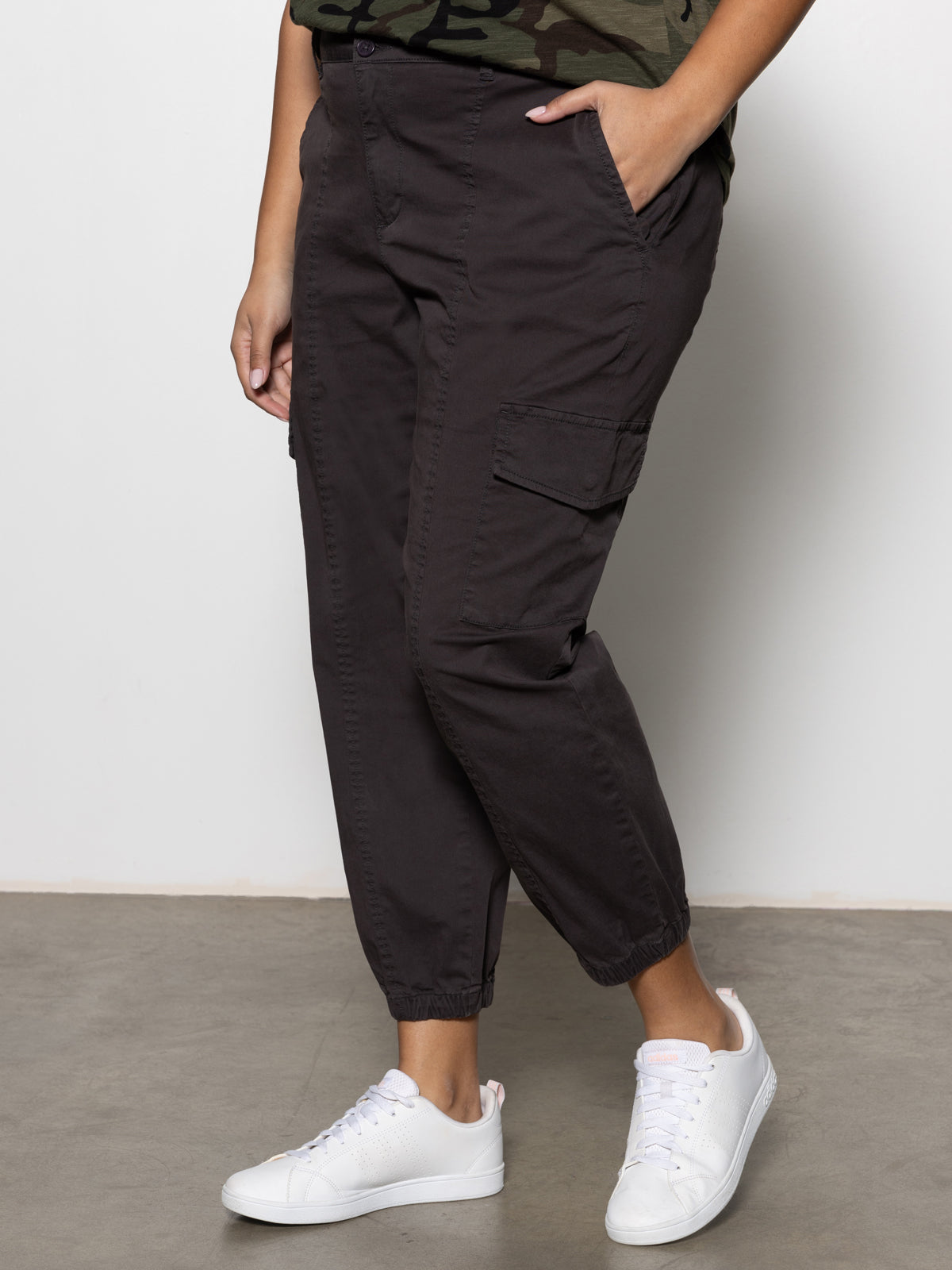 A person wears Sanctuary Clothing's rebel pant obsidian from the inclusive collection, paired with a green top and white sneakers. They stand against a plain wall, hands in pockets; only their lower body is visible.