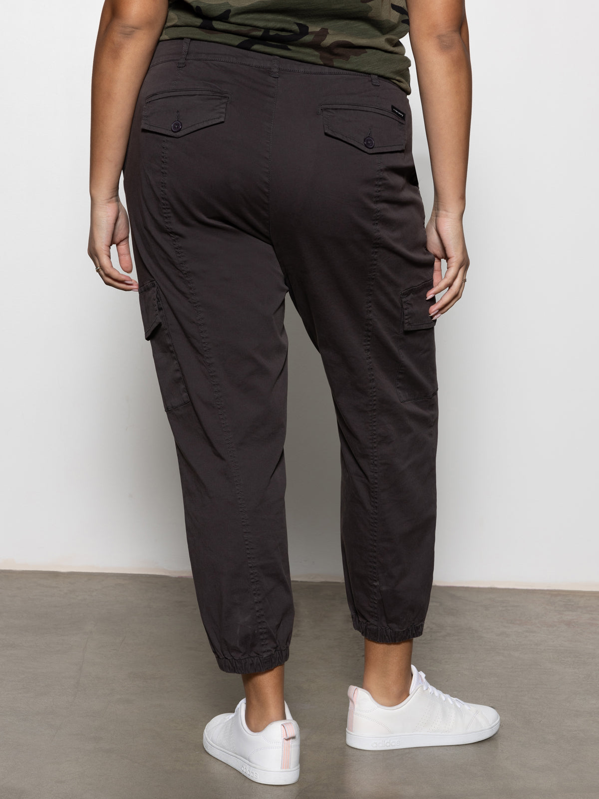 A person faces away, wearing Sanctuary Clothing's rebel pant obsidian from the inclusive collection, styled with elastic cuffs, white sneakers, and a green camo shirt. The setting features a plain concrete floor and a white wall.