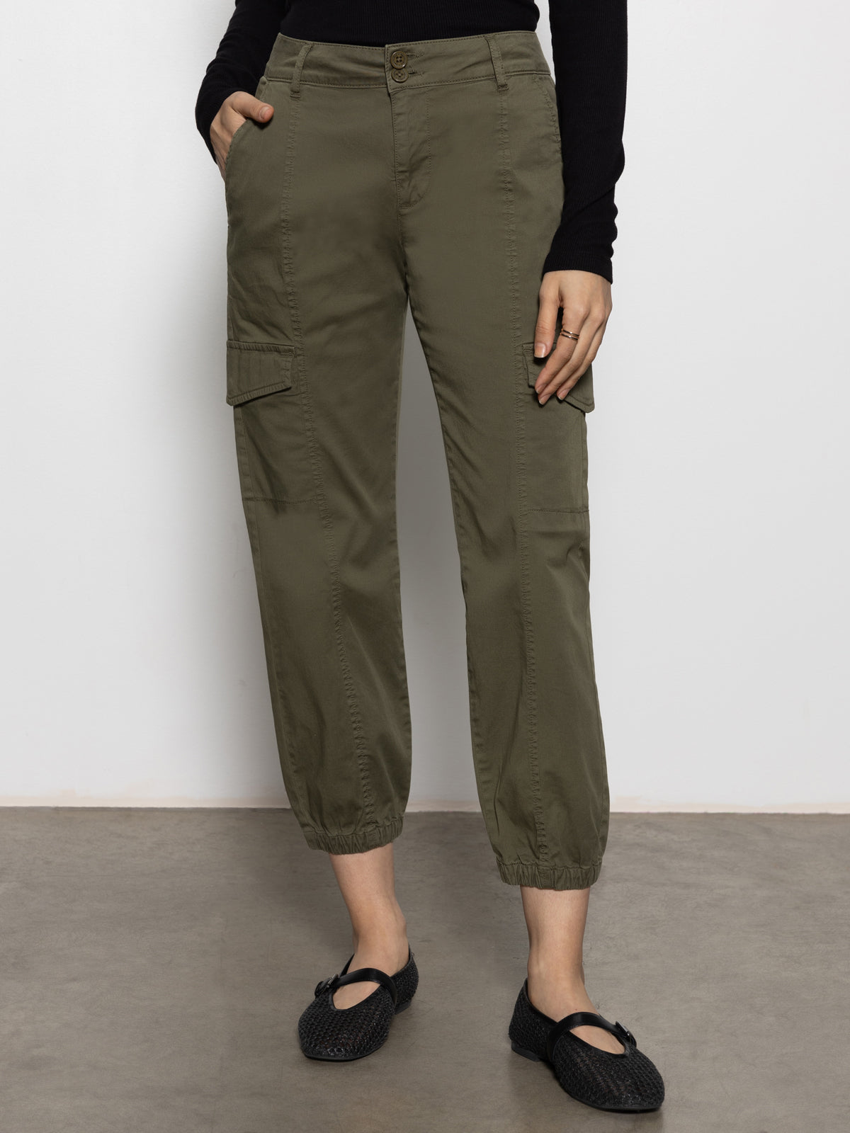 A person models the Sanctuary Clothing rebel pant hiker green cargo joggers, featuring side pockets, paired with black flat shoes; photo shows only the lower half of their body against a plain background.