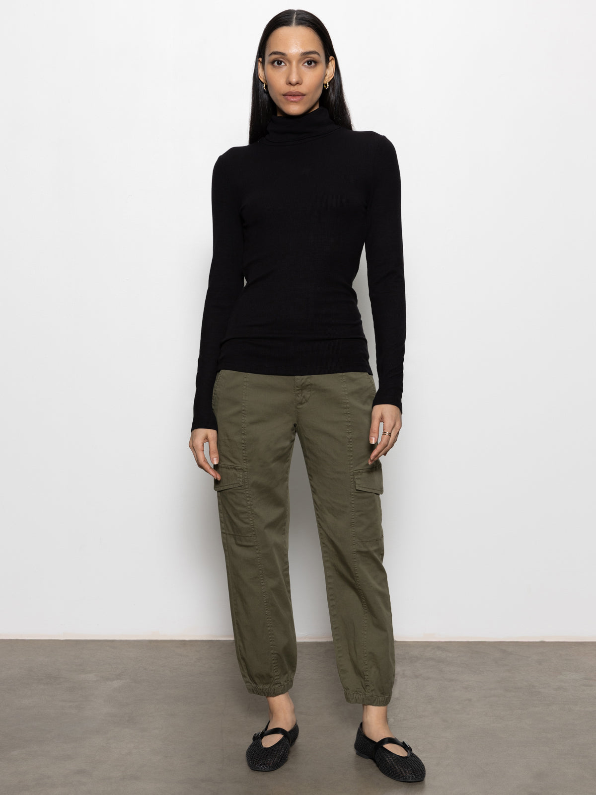 A person with long, straight dark hair and a neutral expression stands against a plain white wall, wearing a black turtleneck, black woven shoes, and the Sanctuary Clothing rebel pant in hiker green.