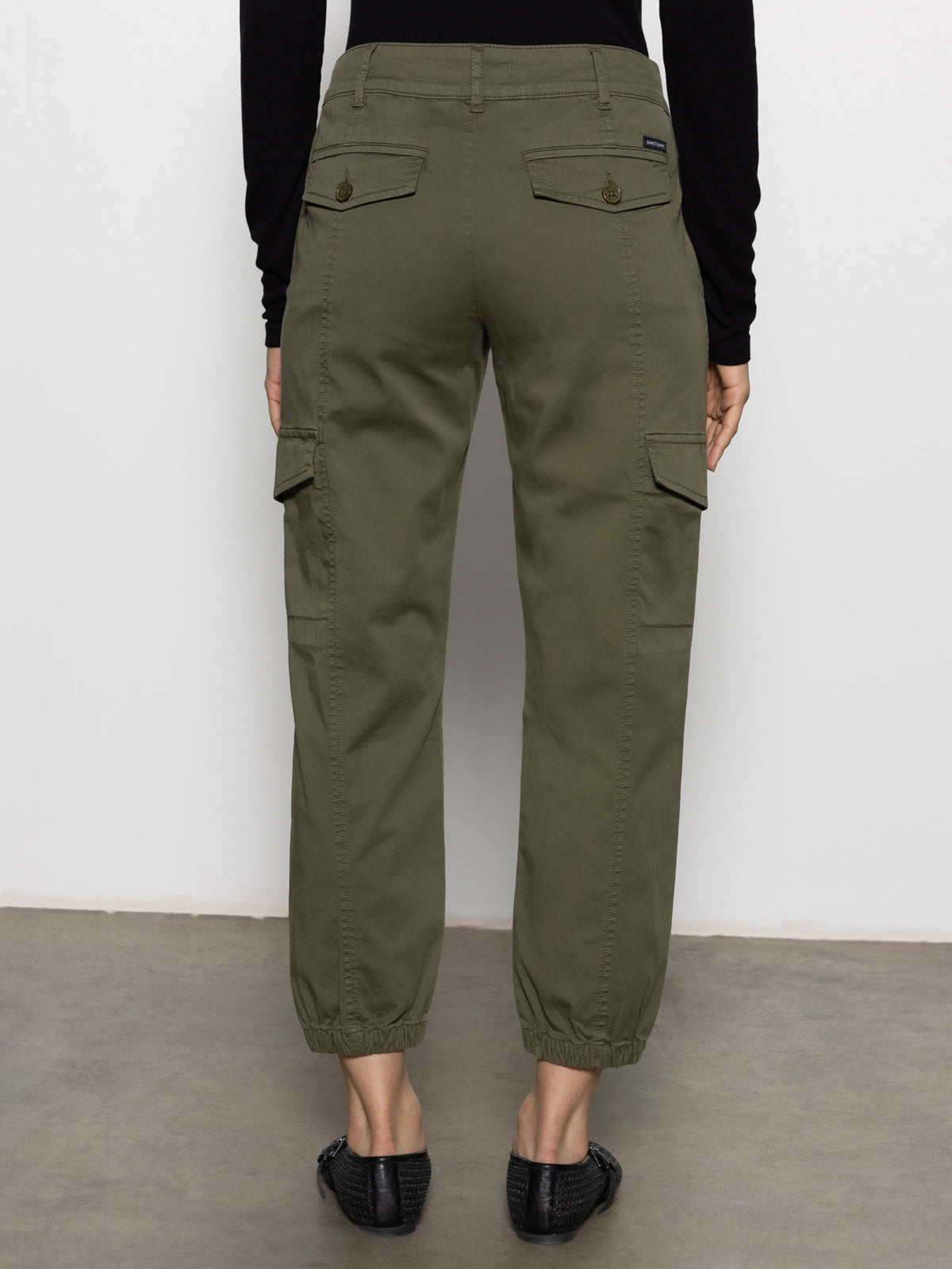 A person wearing Sanctuary Clothing™s rebel pant in hiker green with elastic cuffs and black woven shoes stands facing away from the camera against a plain background.