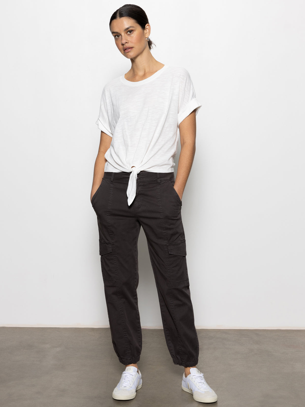A woman stands against a white wall, wearing a white short-sleeve knotted tee, Sanctuary Clothing's rebel pant in obsidian, and white sneakers, with her hands in her pockets and a relaxed expression.