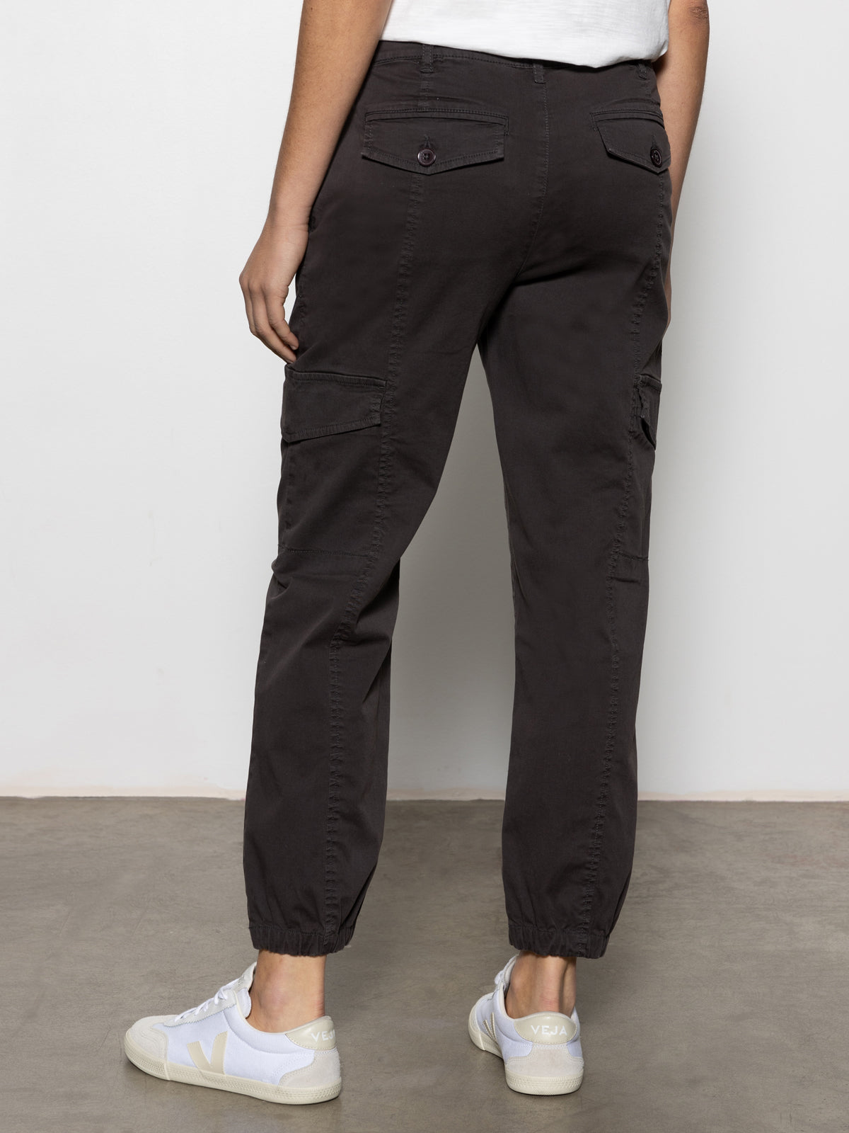 A person faces away indoors, wearing Sanctuary Clothing™s rebel pant obsidian”dark cargo pants with buttoned pockets, cinched ankles, and white sneakers”against a plain backdrop with a gray floor and white wall.