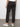 A person faces away indoors, wearing Sanctuary Clothing™s rebel pant obsidian”dark cargo pants with buttoned pockets, cinched ankles, and white sneakers”against a plain backdrop with a gray floor and white wall.