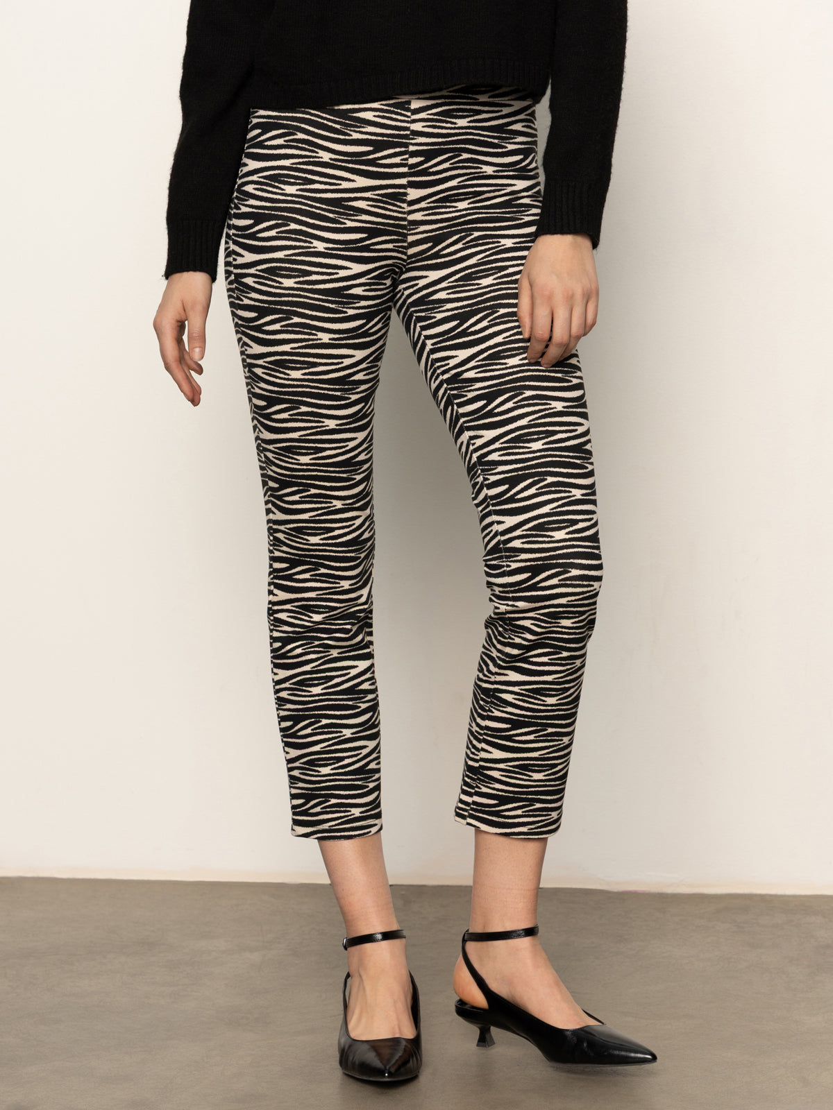 A person wears Sanctuary Clothing's Carnaby Kick Crop Legging Fluid Zebra pants with a black long-sleeve top and black low-heeled shoes, standing against a plain light background.