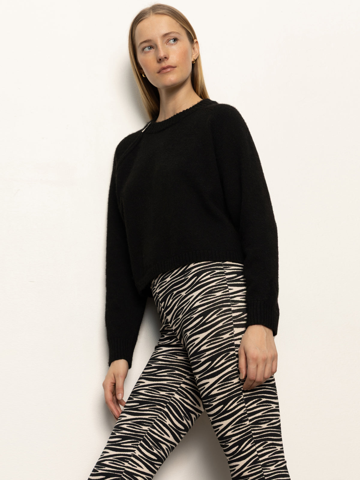A woman with straight blonde hair wears Sanctuary Clothing¢€š¬€ž¢s carnaby kick crop legging fluid zebra pants and a black long-sleeve sweater while standing against a plain white wall.