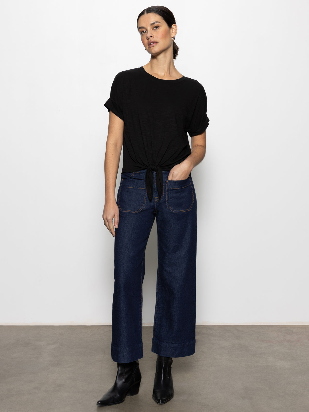 A woman with short dark hair wears Sanctuary Clothing's "the marine magnetic"”a black short-sleeve front-tie top”paired with dark wide-leg jeans and black ankle boots, standing against a plain white wall with her head slightly tilted.