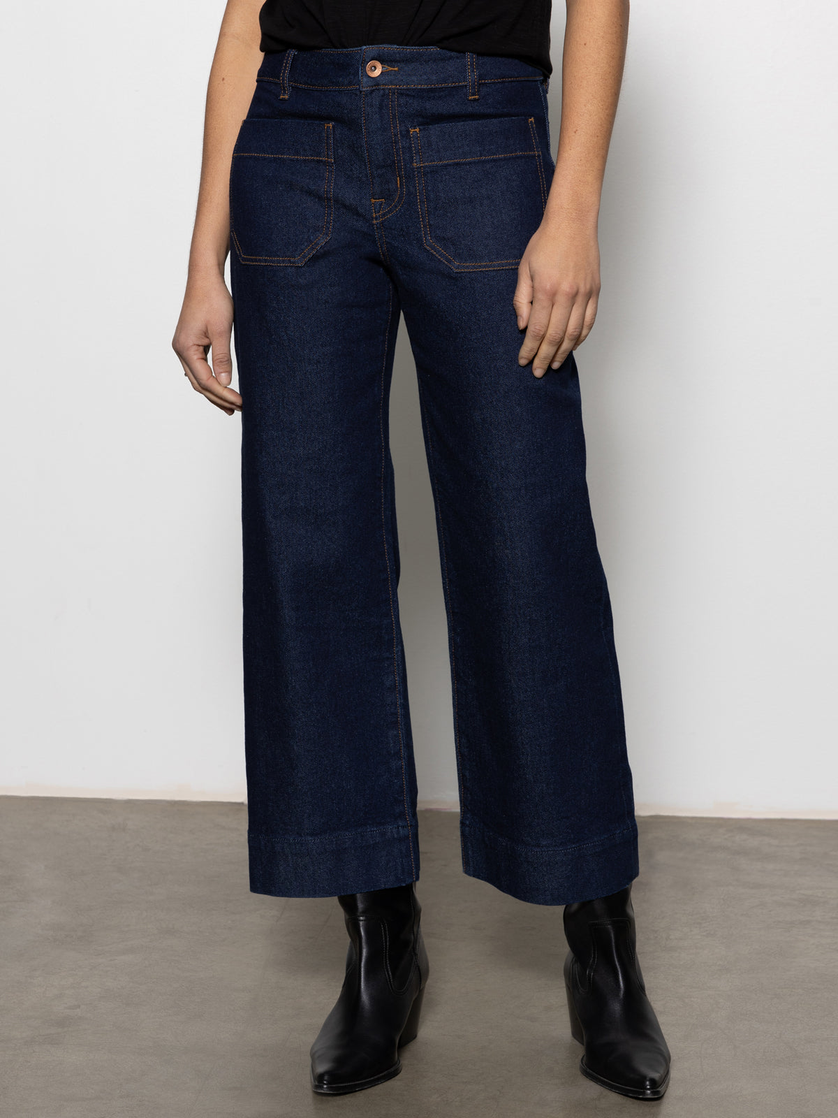 A person wears Sanctuary Clothing's the marine magnetic wide-leg, dark blue jeans with front patch pockets, paired with a black top and black ankle boots, standing against a light-colored wall and concrete floor.