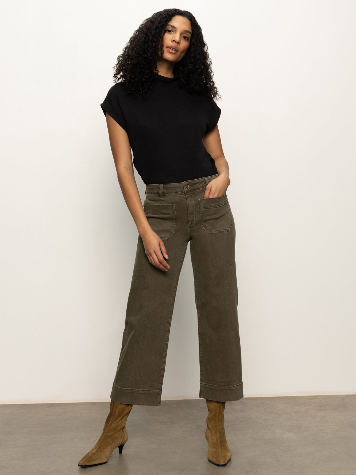 A woman with curly hair wears the Sanctuary Clothing marine crop trouser pant fatigue in olive green, a black short-sleeve top, and brown suede pointed-toe boots, standing against a plain off-white background.