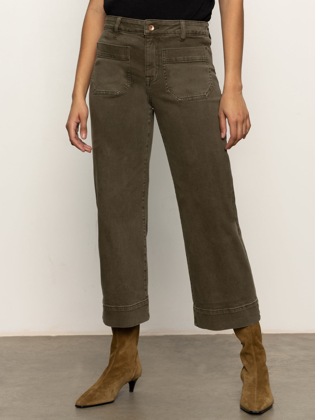 A person wearing Sanctuary Clothing™s marine crop trouser pant fatigue in olive green with front patch pockets, paired with a black top and brown suede heeled ankle boots, stands on a gray floor against a plain light background.