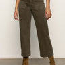 A person wearing Sanctuary Clothing™s marine crop trouser pant fatigue in olive green with front patch pockets, paired with a black top and brown suede heeled ankle boots, stands on a gray floor against a plain light background.