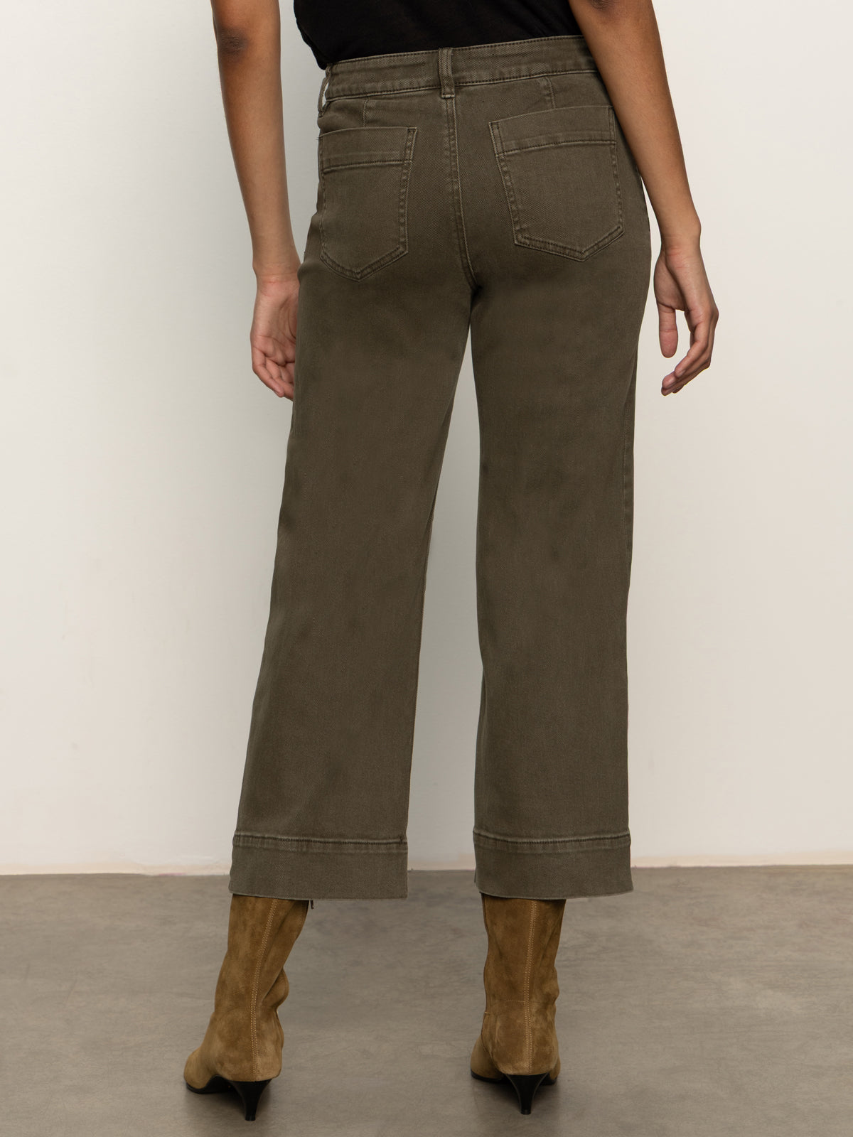 A person stands facing away, wearing Sanctuary Clothing's the marine crop trouser pant fatigue”high-waisted, olive green wide-leg jeans with cuffed hems”and brown heeled boots against a plain background.