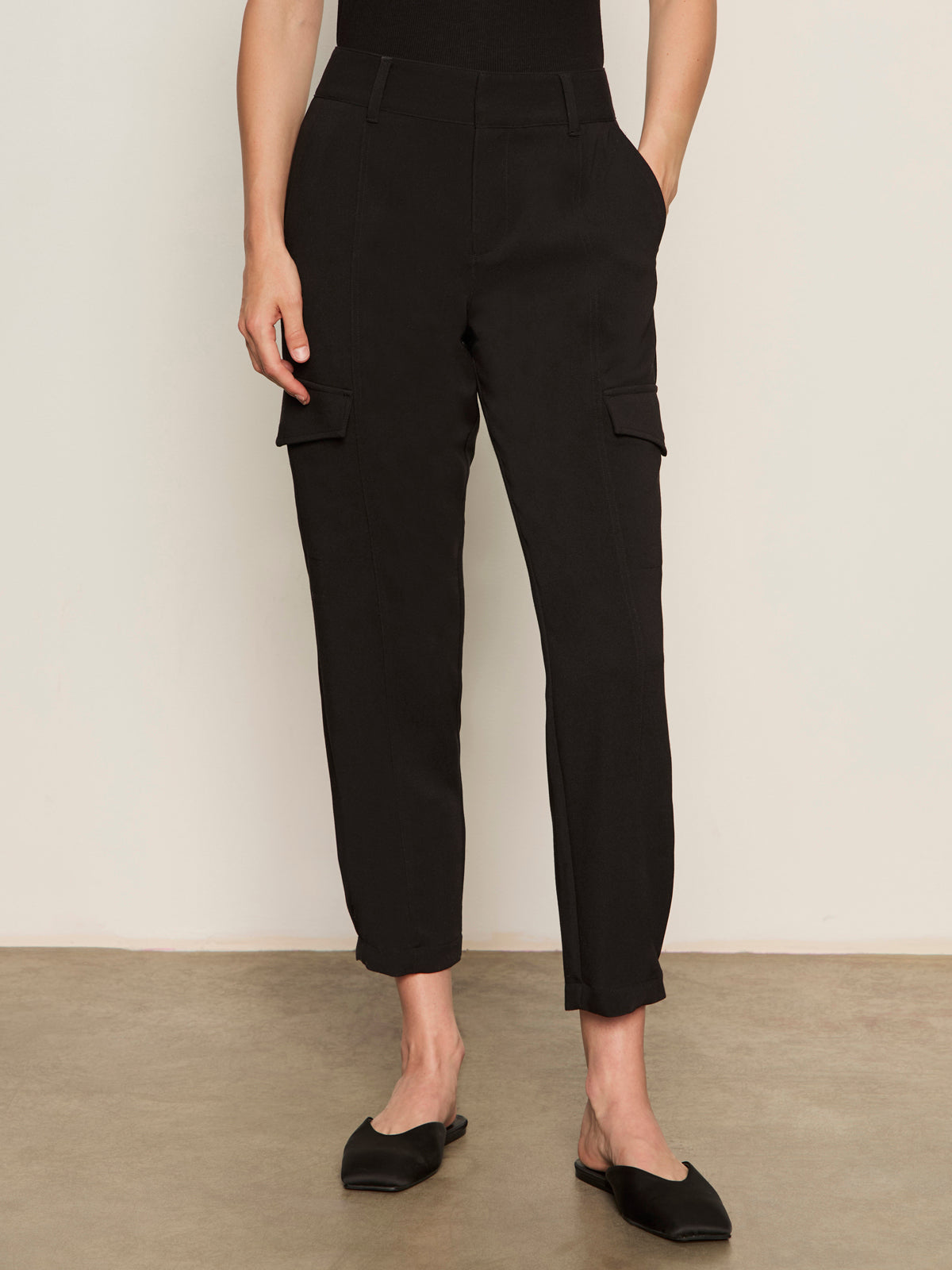 A person wears Sanctuary Clothing's polished cargo pant in black, featuring side pockets and a cropped fit, standing on a brown floor against an off-white wall with black slip-on shoes.