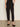 A person wears Sanctuary Clothing's polished cargo pant in black, featuring side pockets and a cropped fit, standing on a brown floor against an off-white wall with black slip-on shoes.