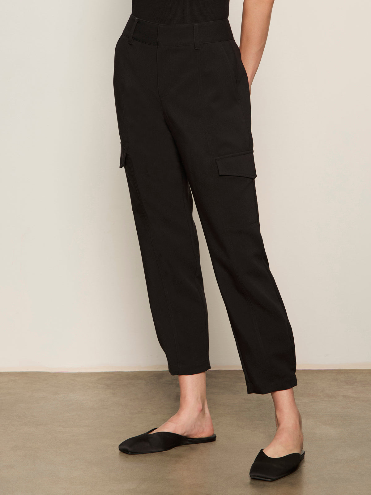 A person wears Sanctuary Clothing's polished cargo pant in black with black slip-on shoes, standing against a plain light background and posing with one hand in their pocket.