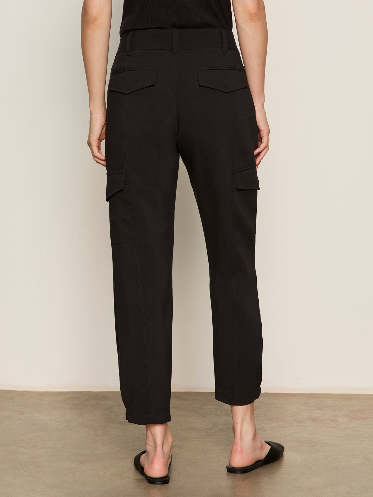 A person stands facing away in Sanctuary Clothing's polished cargo pant black, featuring back pockets and a straight-leg fit, paired with black flat shoes against a neutral background.