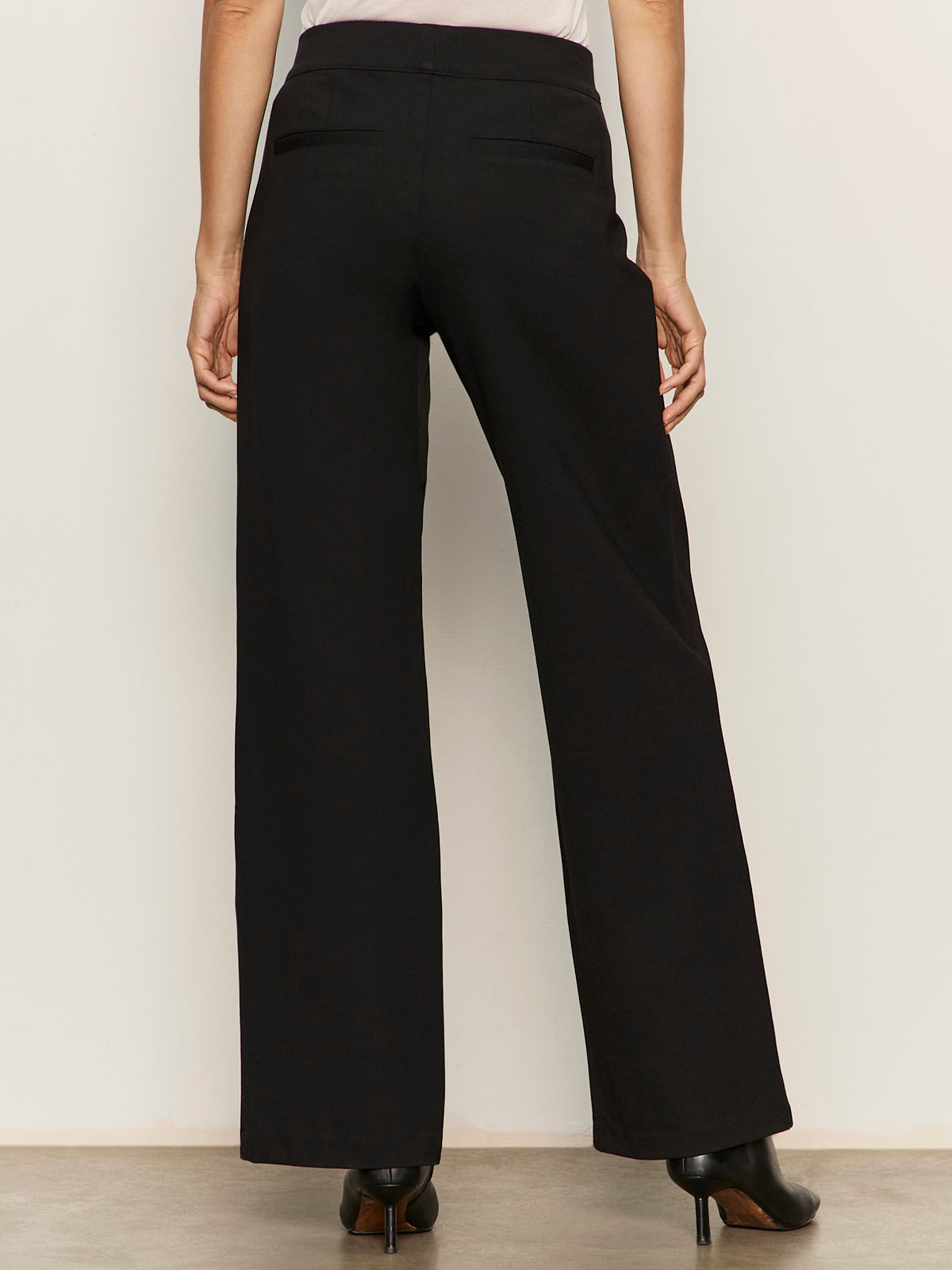 A person faces away wearing the Sanctuary Clothing runway ponte trouser in black�high-waisted, wide-leg pants with two back welt pockets, paired with black heeled shoes. The look is set against a plain, light-colored background.