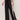 A model wears Sanctuary Clothing’s black casual track pant summer sand with a white side stripe, paired with a black top and brown flat sandals, standing against a plain light background.