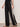 A model wears Sanctuary Clothing’s black casual track pant summer sand with a white side stripe, paired with a black top and brown flat sandals, standing against a plain light background.