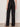 A person stands indoors from the waist down, wearing Sanctuary Clothing's casual track pant black summer sand—loose black pants with a white side stripe, elastic waistband, and flat sandals. The light background is plain.