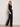 A woman in Sanctuary Clothing's casual track pant black summer sand poses confidently against a plain light wall, pairing the pants with sunglasses and a sleeveless black buttoned top.
