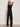 A woman in Sanctuary Clothing's casual track pant black summer sand pairs it with a sleeveless black top and sunglasses, standing against a plain light background with one arm bent and her hand touching her head.
