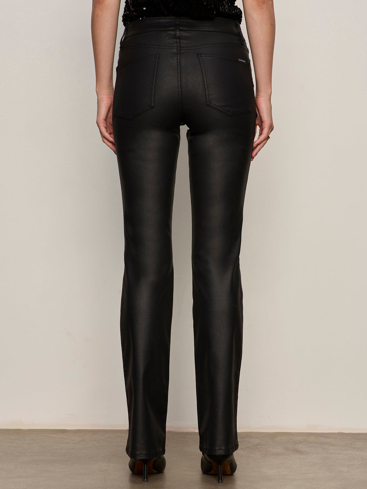 A person faces away from the camera, wearing Sanctuary Clothing's slick brook bootcut pant in black€�high-waisted, straight-leg, shiny material€�paired with dark heeled shoes against a plain neutral background.