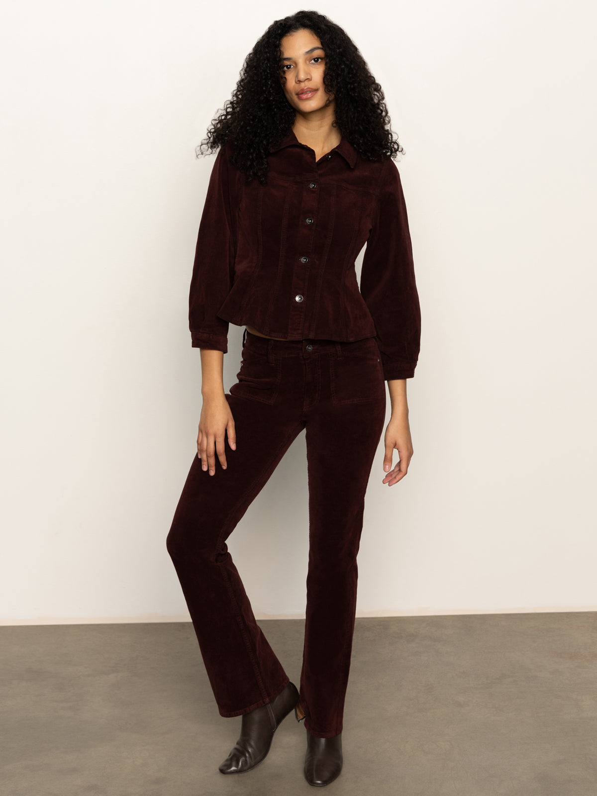 A woman with curly hair wears Sanctuary Clothing„¢s velveteen brook bootcut pant in dark crimson, paired with a matching jacket and brown ankle boots, standing against a plain light background.