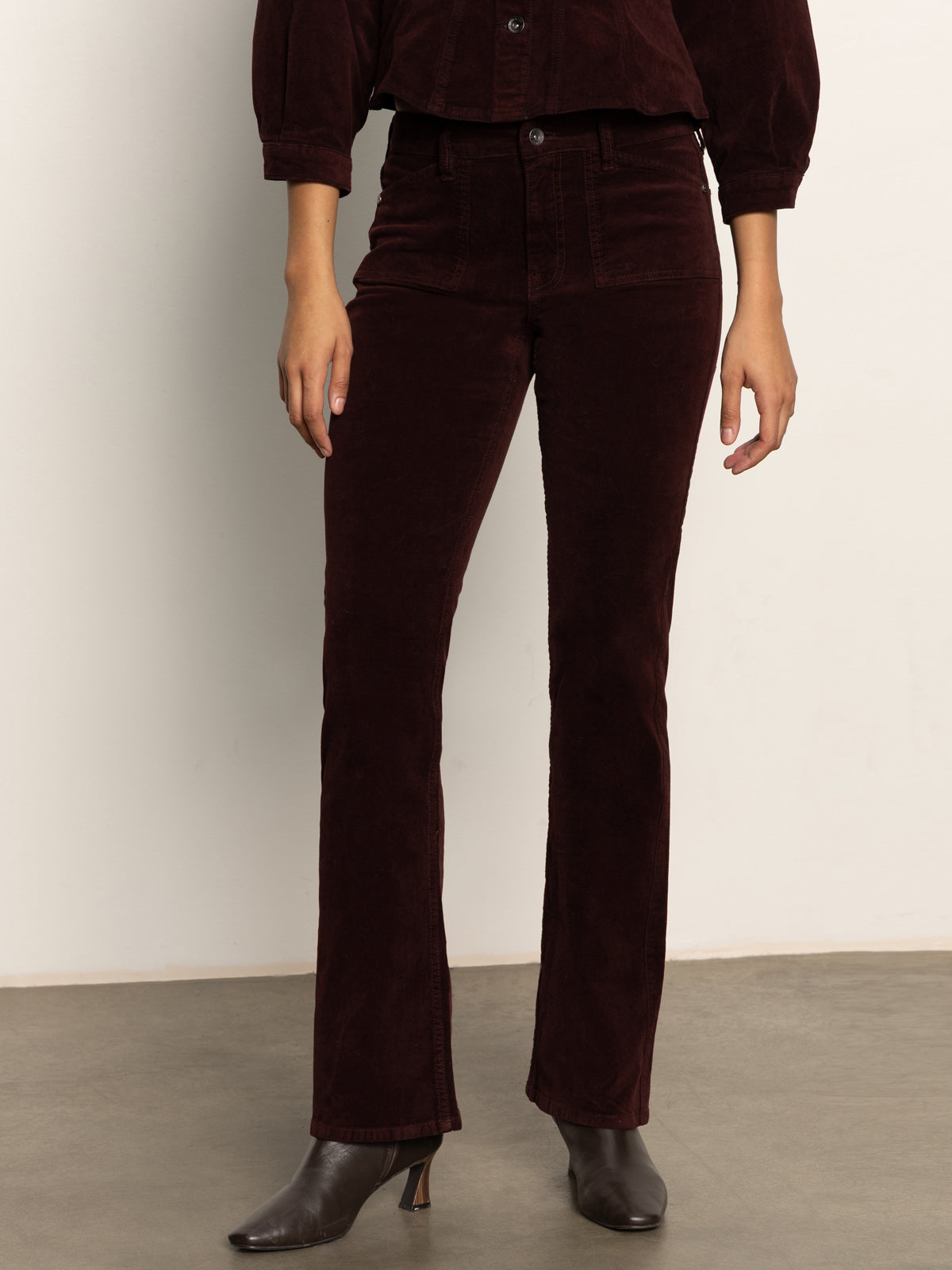 A person indoors wears Sanctuary Clothing„¢s velveteen brook bootcut pant in dark crimson, paired with matching outerwear and heeled ankle boots. Only their lower half is visible against a plain background.