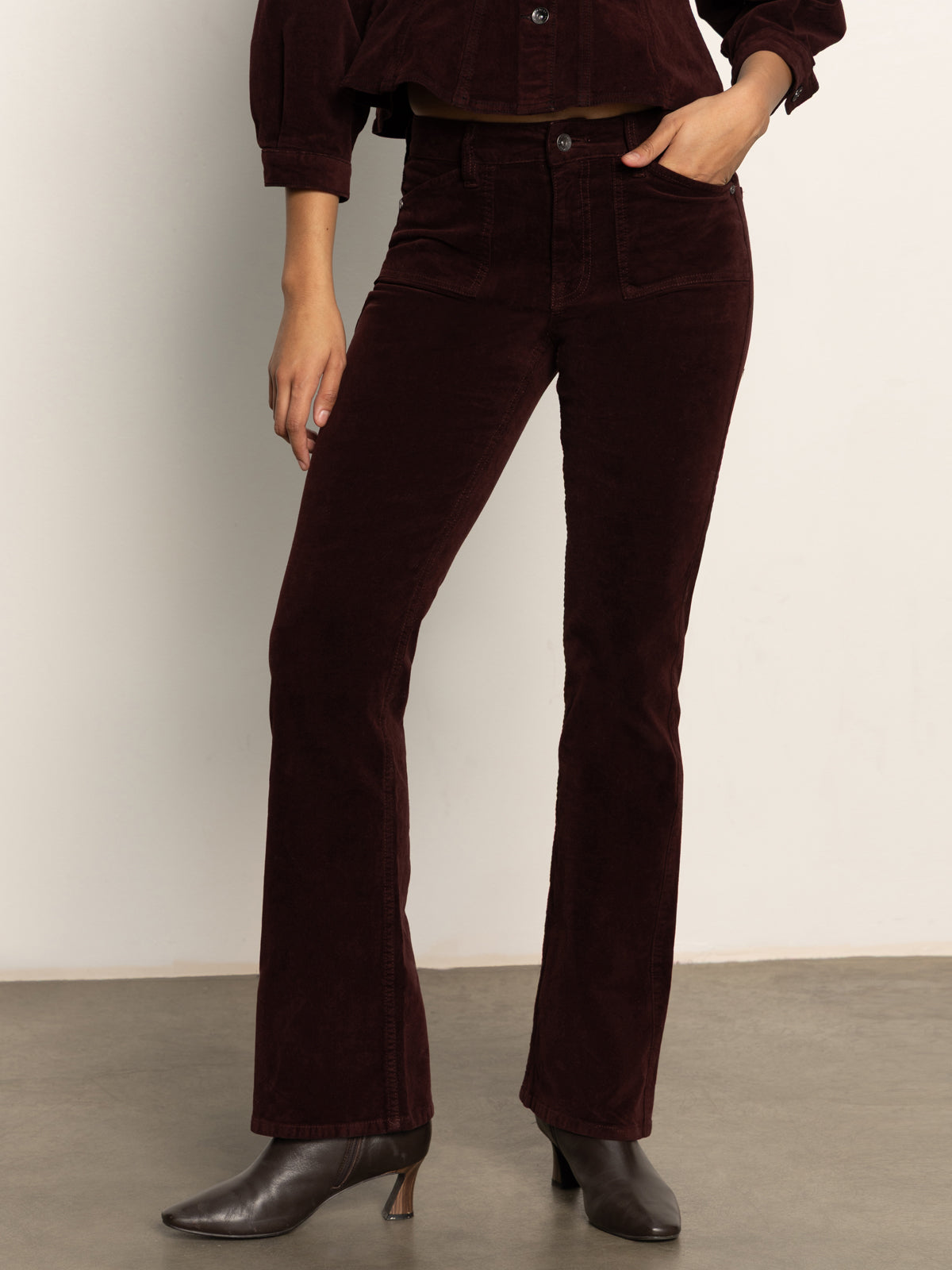 A person wearing Sanctuary Clothing's velveteen brook bootcut pant in dark crimson stands against a plain light background. The photo is cropped to show the body from shoulder to foot.