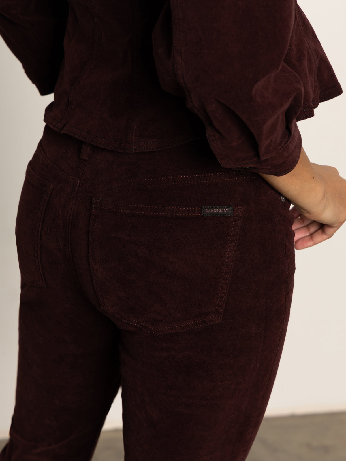 A person shown from the waist down and viewed from behind wears Sanctuary Clothing„¢s velveteen brook bootcut pant in dark crimson, paired with a matching top. Their right arm rests by their side against a neutral background.