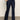 A person models the Sanctuary Clothing denim stunner jean plot twist, showcasing its high-waisted, dark blue design paired with black heeled boots and one hand in their pocket; only the lower body is visible against a plain background.