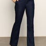 A person models the Sanctuary Clothing denim stunner jean plot twist, showcasing its high-waisted, dark blue design paired with black heeled boots and one hand in their pocket; only the lower body is visible against a plain background.