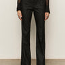 A person wears Sanctuary Clothing's the stunner pant luster stripe€�a black, high-waisted, straight-leg pant with subtle glittery pinstripes€�paired with a sheer black top and black shoes, standing against a plain light background.