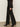 A person wears the Sanctuary Clothing "the stunner pant luster stripe" black, pinstriped wide-leg trousers, paired with a sheer black shirt and black heeled boots indoors on a light-colored floor; only the lower half is visible.