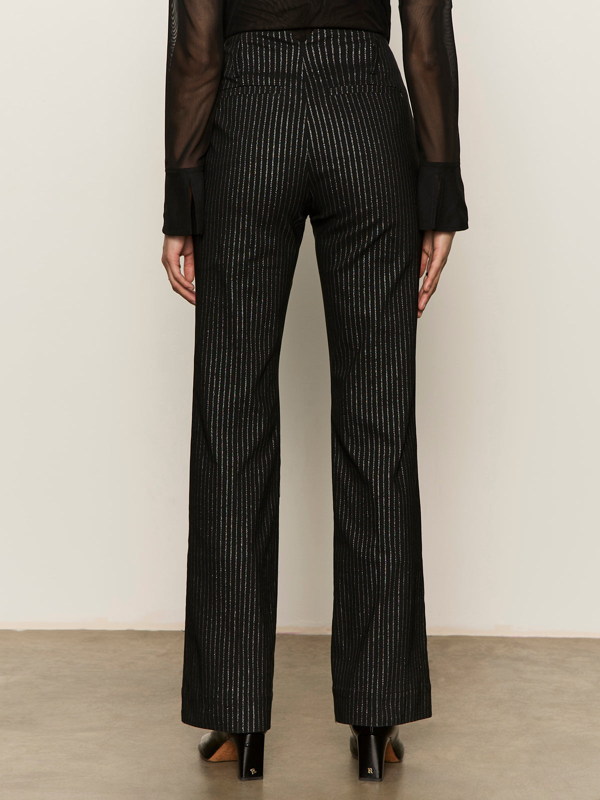 A person stands with their back turned, wearing Sanctuary Clothing's the stunner pant luster stripe€�black high-waisted pants with vertical white pinstripes€�paired with a sheer black top and black shoes against a plain background.