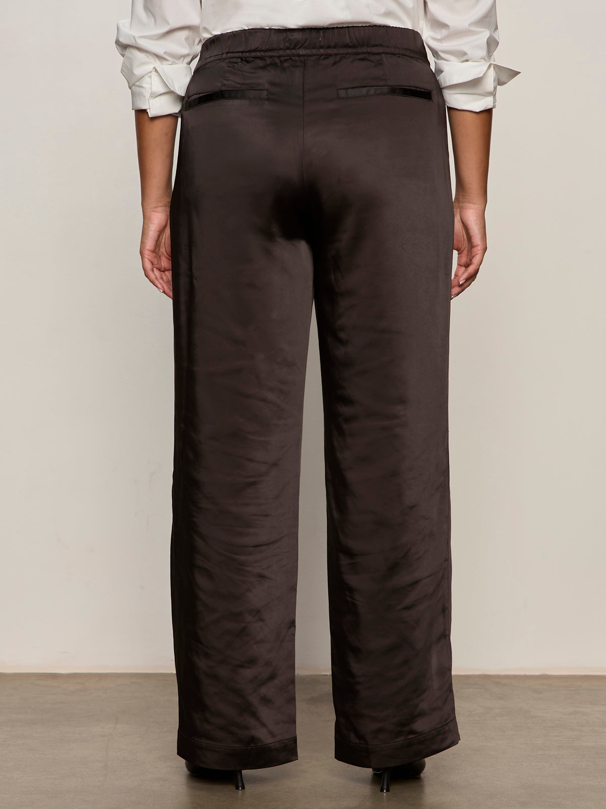 A person stands facing away, wearing Sanctuary Clothing's modern satin trouser in black (extended sizing) and a white shirt with rolled-up sleeves. The trousers feature welt back pockets, and the person stands on a neutral-colored floor.