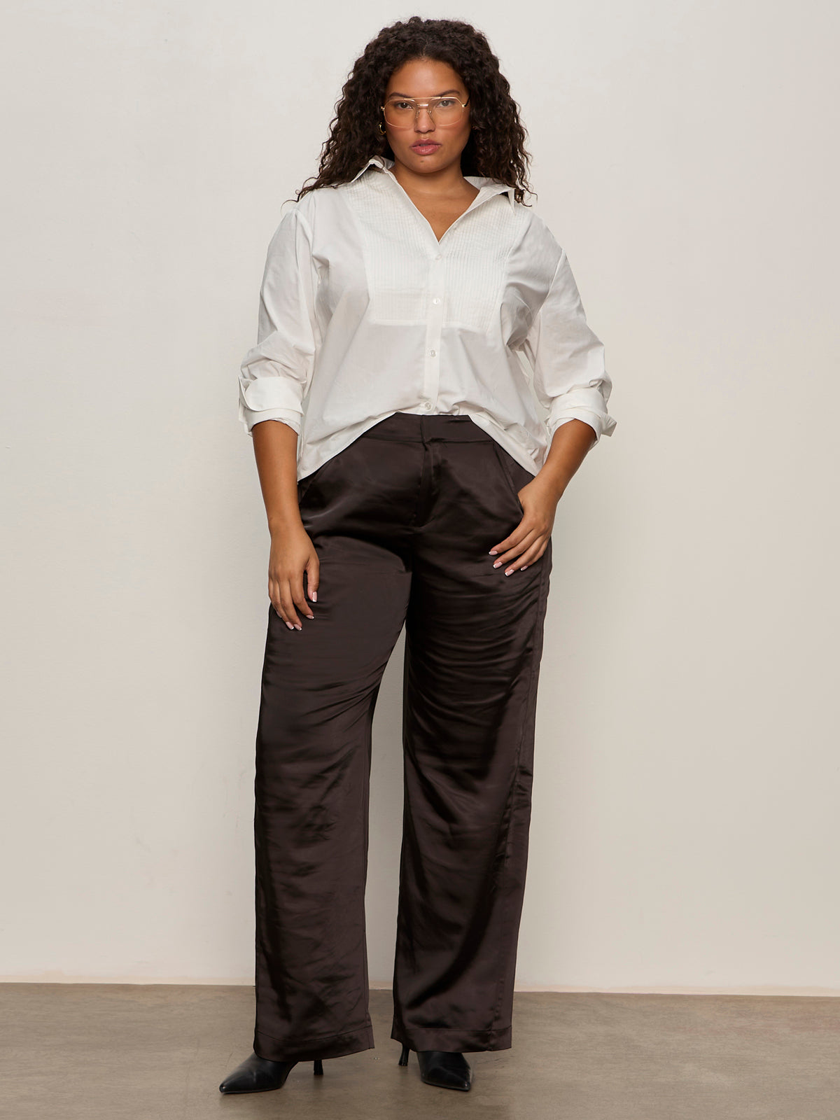 A woman wearing glasses and a white button-up shirt stands against a light background, styled in Sanctuary Clothing's modern satin trouser black extended sizing and black pointed-toe shoes.