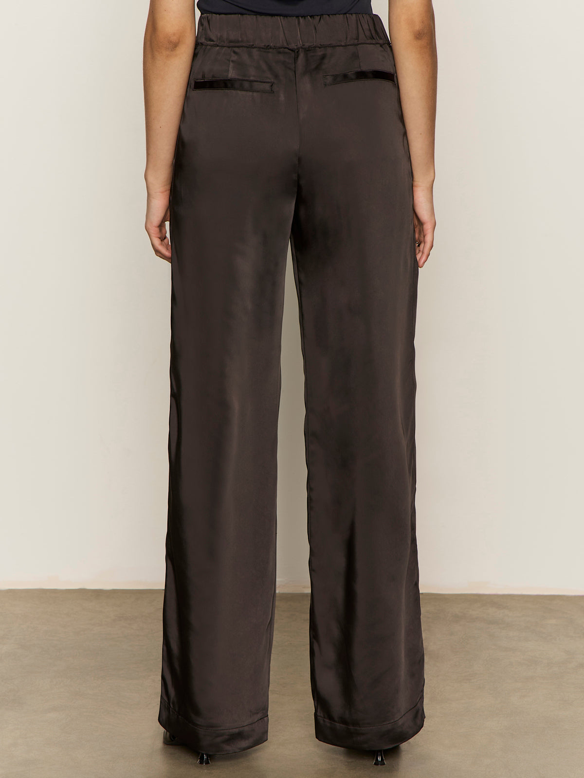 A person faces away from the camera, wearing Sanctuary Clothing„¢s modern satin trouser in black, featuring back pockets, against a plain light wall and concrete floor.