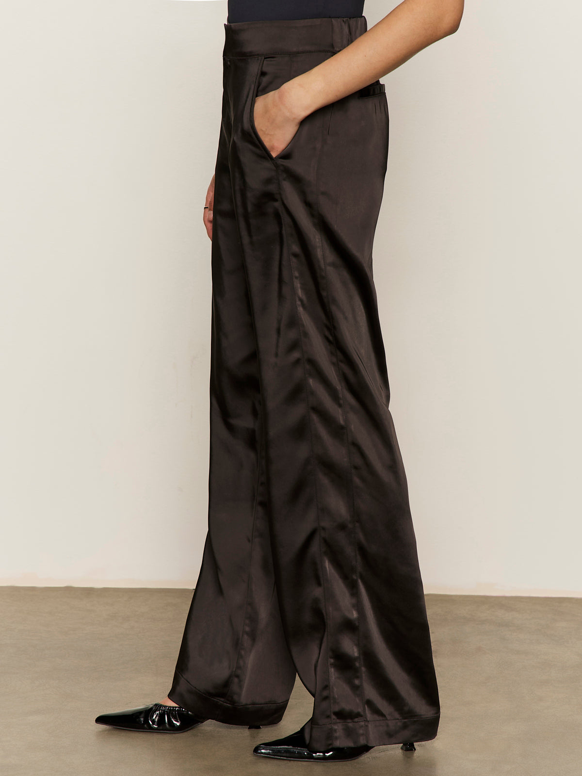 A person models the modern satin trouser black from Sanctuary Clothing, styled with a black top and pointed shoes, standing sideways with one hand in the pocket against a plain background.