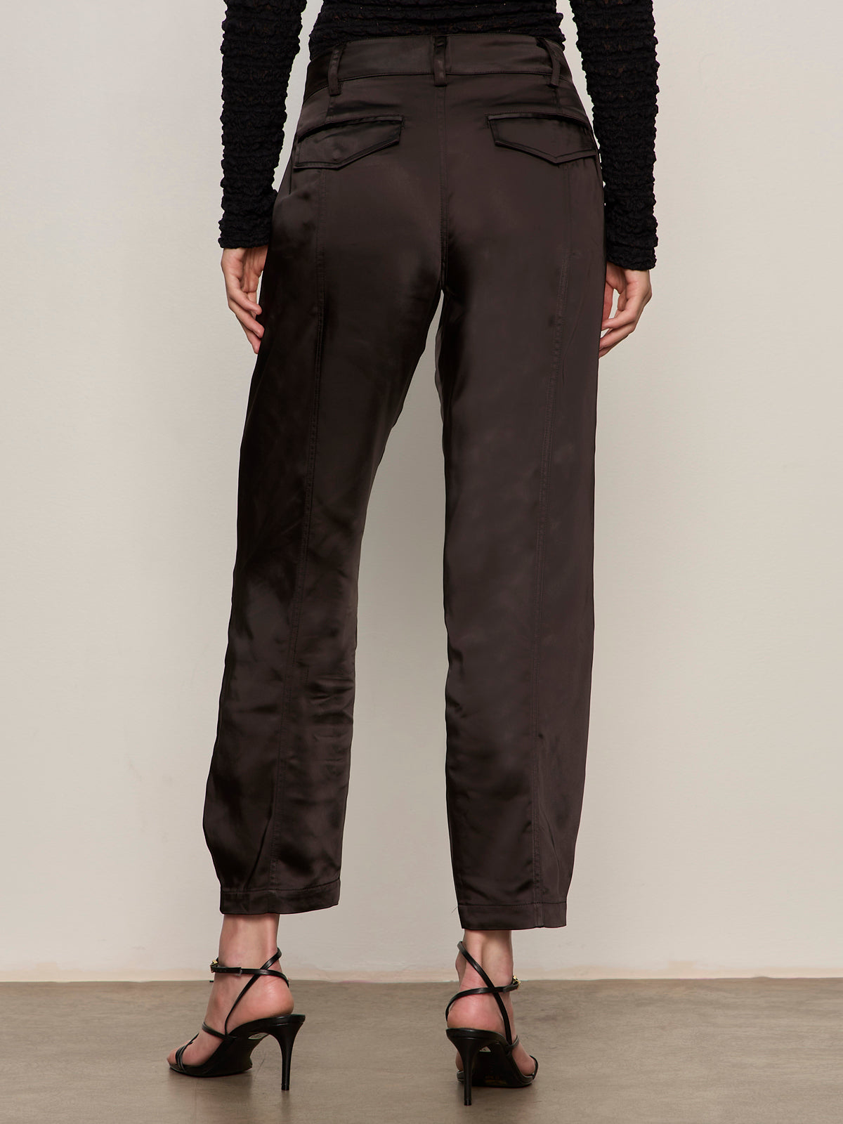 A person stands facing away from the camera wearing Sanctuary Clothing's Sahara Satin Tapered Pant in black, paired with a textured black long-sleeve top and black strappy heeled sandals.