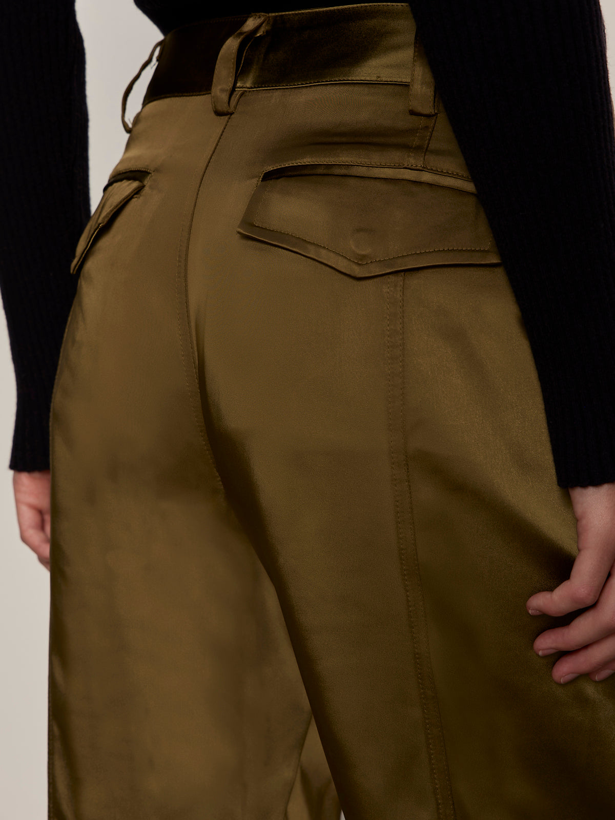Close-up of Sanctuary Clothing's sahara satin tapered pant in Dirty Martini, featuring shiny olive green fabric and back flap pockets. Pants are styled with a black ribbed long-sleeve top, shown from waist to mid-thigh.