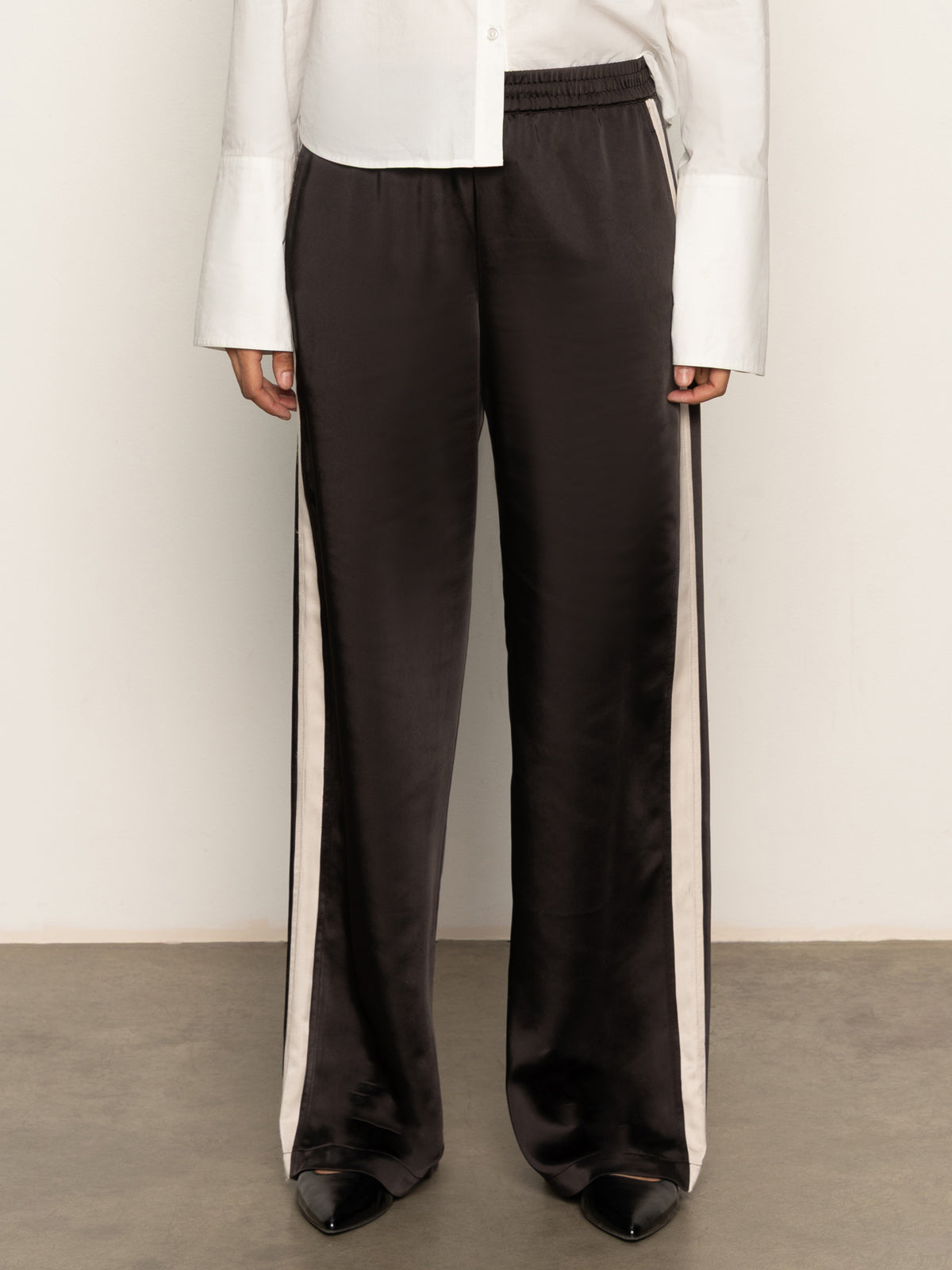 A person wears Sanctuary Clothing„¢s satin track pant in black with white side stripes, a white long-sleeve shirt with wide cuffs, and black pointed-toe shoes, standing against a plain light background.